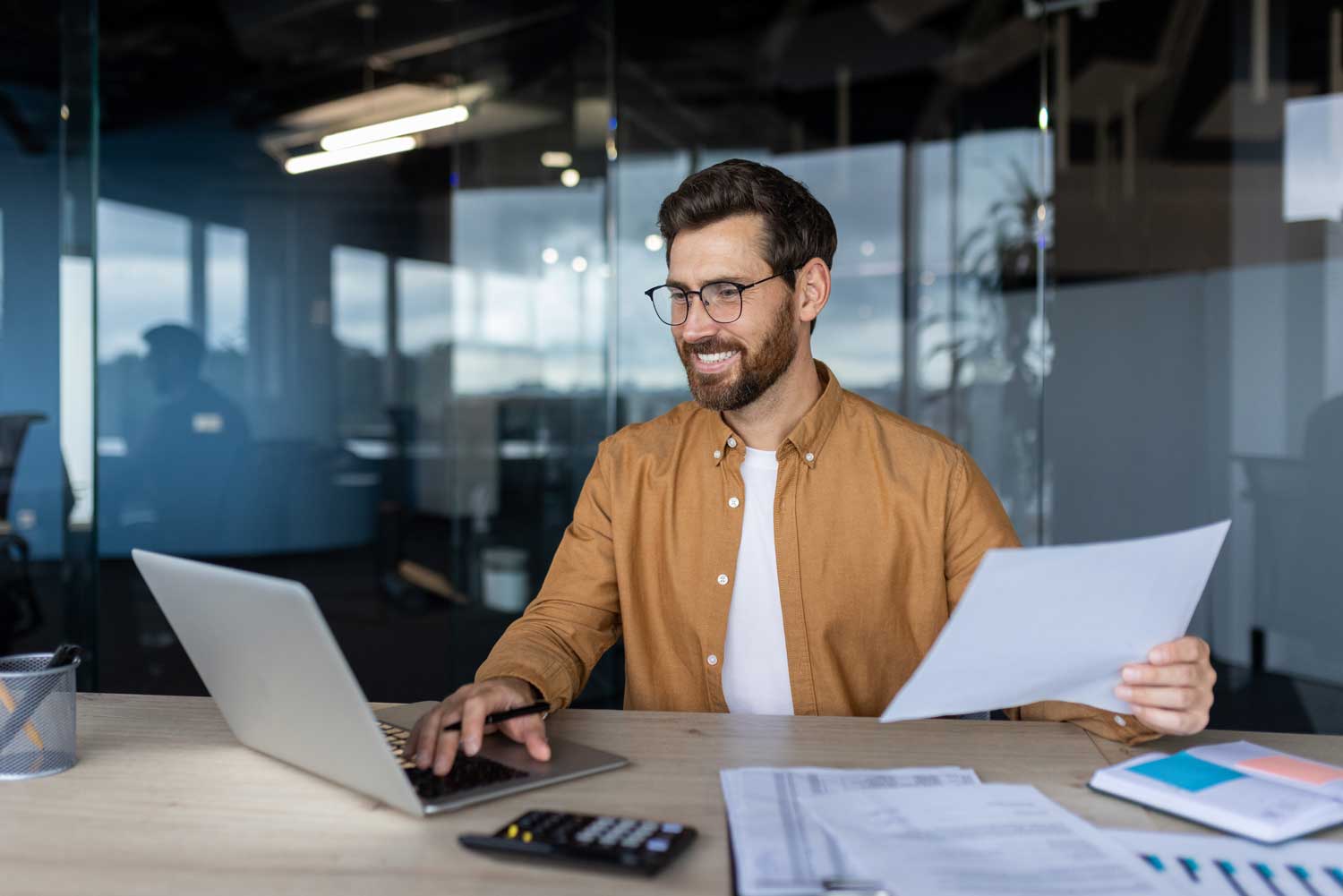 Man smiling while typing on his laptop and holding a piece of paper.