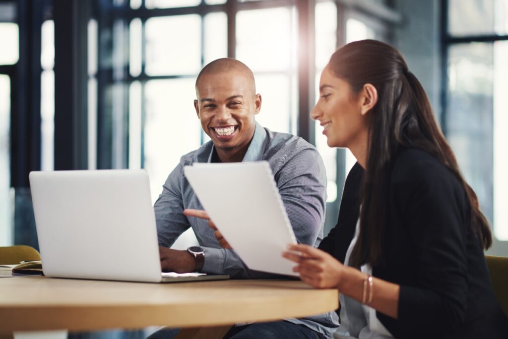 Man smiling at the camera while typing into a laptop, woman holding a piece of paper and pointing at something out of frame.