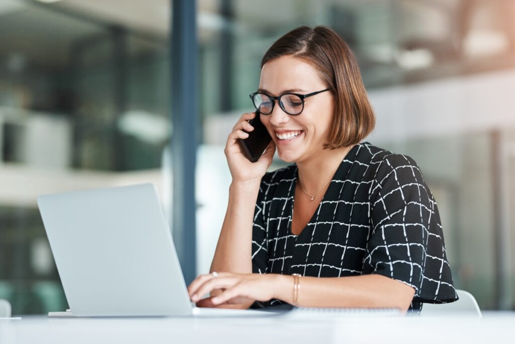 Woman smiling while holding her cellphone and typing into her laptop.