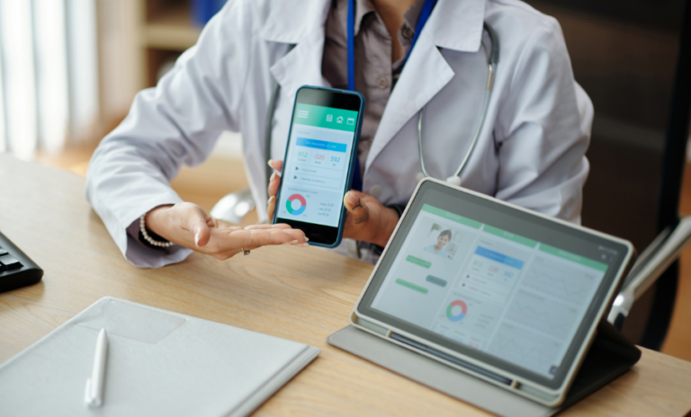 A person in a white lab coat is holding a smartphone and laptop