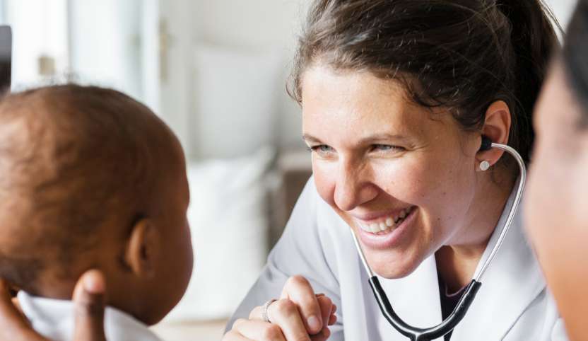 A woman doctor wearing a stethoscope, holding a baby's hand.